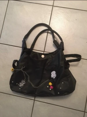 Svart handväska med kedjedetaljer - Cool svart handväska i syntetmaterial med dubbla handtag och axelrem. Väskan har dekorativa kedjor, nitar, en vit blomma och färgglada pärlor framtill. Baksidan har en praktisk dragkedjeficka. Perfekt för dig som gillar unika detaljer! Har tre fack, samt ficka på baksidan.