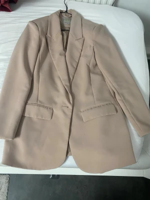Beige kavaj från Aware - Stilren beige kavaj från Aware med klassisk passform och markerade axlar. Kavajen har två fickor med lock framtill och är helfodrad. Perfekt för en trendig och clean look. Materialet känns mjukt och har en lätt glans. Sista bilden visar en blå fläck! 