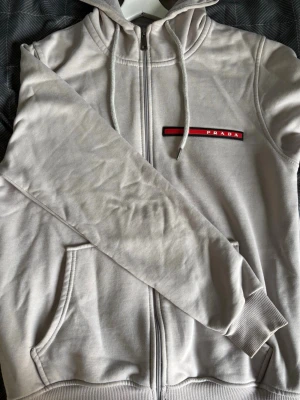 Beige hoodie med dragkedja från Prada - Snygg beige hoodie med dragkedja från Prada. Hoodien har huva med dragsko, fickor framtill och en röd logodetalj på bröstet. Tillverkad i mjukt material som känns skönt mot huden. Perfekt för en avslappnad och trendig look.