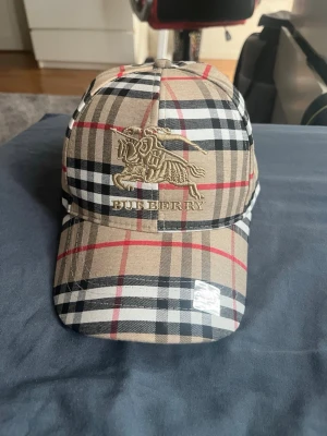Burberry beige rutig keps - Snygg keps från Burberry i klassiskt beige, svart, vitt och rött rutmönster. Framtill finns ett broderat Burberry-logotyp med riddare. Kepsen har böjd skärm och justerbar baksida. Tillverkad i bomull för en bekväm känsla.