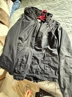 Svart vindjacka från H&M med huva - Snygg svart vindjacka från H&M med dragkedja framtill och två fickor med lock. Jackan har en praktisk huva och resår i nederkant för bättre passform. Perfekt för blåsiga dagar och enkel att matcha med olika outfits.