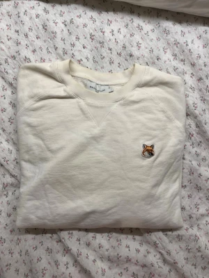 Vit sweatshirt från Maison Kitsuné - Säljer en stilren vit sweatshirt från Maison Kitsuné med den ikoniska rävpatchen på bröstet. Tröjan har rund halsringning och ribbade muddar vid ärmslut och nederkant. Perfekt för en clean och avslappnad look. Använd fåtal gånger.