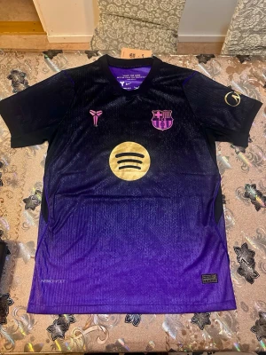 Barcelona away 26/27 Lamine Yamal #10  - Snygg Barcelona matchtröja player version i svart och lila med guldiga detaljer och tryck. Tröjan har Lamine Yamal och nummer 10 på ryggen, samt klubbmärke och sponsorlogga på bröstet. Tillverkad i ett lätt och ventilerande material med Aero-Fit teknologi. 