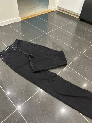 Svarta straight fit jeans - Säljer ett par svarta jeans med rak passform och klassisk femficksdesign. Jeansen har knappgylf och nitar vid fickorna för extra detaljer. Perfekta för dig som gillar en enkel och stilren look. Som 36 i midjan och 32 i längden
