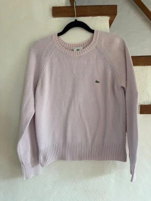 Ljusrosa stickad tröja från Lacoste - Supersnygg ljusrosa stickad tröja från Lacoste med klassisk rund halsringning och ribbade muddar. Tröjan har den ikoniska gröna krokodilloggan broderad på bröstet och är perfekt för dig som gillar stilrena plagg med en sportig touch.