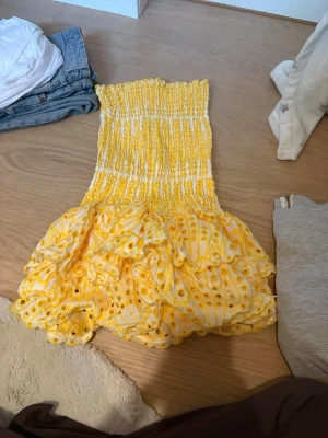Gul volangklänning från Designbyamor - Supersöt kort gul klänning från Designbyamor med smockad överdel och volangig kjol. Klänningen har broderade detaljer och är perfekt för sommarens alla tillfällen. Tillverkad i bomull för en luftig och bekväm känsla. Använd 1 gång.