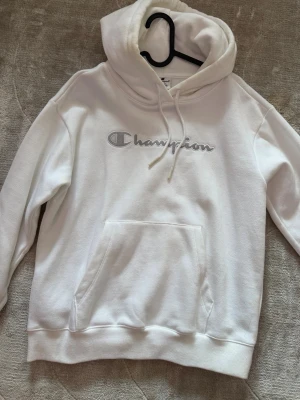 Vit hoodie från Champion - Snygg vit hoodie från Champion med broderad logga i silvergrått på bröstet. Klassisk känguruficka framtill, ribbade muddar och justerbar huva med dragsko. Perfekt för en chill och sportig look.