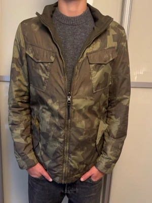 Camouflage vindjacka från Woolrich - Snygg camouflagefärgad vindjacka från Woolrich med dragkedja framtill och två stora bröstfickor. Jackan har hög krage och diskret logga på ärmen. Perfekt för dig som gillar streetwear och vill ha en lätt jacka med coolt mönster.