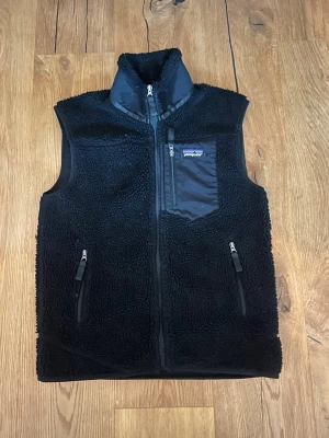 Svart fleeceväst från Patagonia - Mysig svart fleeceväst från Patagonia med hög krage och dragkedja framtill. Västen har två fickor med dragkedja och en bröstficka med logga. Tillverkad i mjukt teddyfleece-material som håller värmen och ger en avslappnad look.
