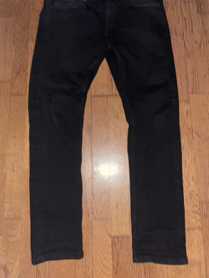 Svarta jeans från SMOG/DENIM - Säljer ett par svarta jeans från SMOG/DENIM i storlek 31/30. Jeansen har en klassisk rak passform och är tillverkade i ett mjukt denimtyg. Perfekta för dig som gillar en clean och enkel stil.