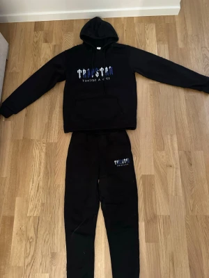 Svart trapstar tracksuit  - Säljer en svart tracksuit med hoodie och matchande byxor. Hoodien har trycket 'TRUST NO ONE IT'S A SECRET' i vitt och blått på bröstet samt en stor ficka framtill. Byxorna har samma tryck på ena benet. Perfekt för chill eller träning.