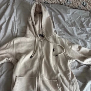 Beige hoodie från Polo Ralph Lauren - Säljer en beige hoodie från Polo Ralph Lauren med dragkedja framtill och klassisk huva med dragsko. Tröjan har det ikoniska Polo-logotypen broderad på bröstet och två fickor framtill. Tillverkad i mjukt material som känns skönt mot huden.