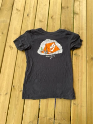 Svart Nike t-shirt med tryck - Svart t-shirt från Nike med ett färgglatt tryck på ryggen som visar ett orange tält med Nike-logga och texten 'Waiting for the drop'. Klassisk passform och korta ärmar, perfekt till vardags för en sportig look.