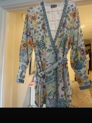 Blommig kortklänning med paljetter  - Färgglad kortklänning med blommigt mönster i vitt, blått, gult, orange och lila. Klänningen har v-ringning, lång ärm, bälte i midjan och är dekorerad med paljetter och broderade detaljer. Perfekt för dig som vill sticka ut med en unik look.