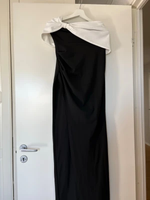 Svart långklänning med vit offshoulder - Elegant svart långklänning med vit offshoulder-detalj och asymmetrisk skärning. Klänningen har en stilren och modern look med dragkedja i ryggen. Perfekt för dig som vill sticka ut med en unik och trendig design.