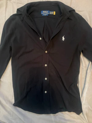 Svart skjorta från Polo Ralph Lauren - Klassisk svart skjorta från Polo Ralph Lauren med vit broderad logga på bröstet. Skjortan har långa ärmar, knappar framtill och en tidlös design som passar till det mesta. Tillverkad i mjuk bomull för en bekväm känsla. Max ändvönd 2-3 gånger. Pris kan diskuteras 