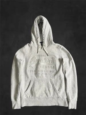 Ljusgrå hoodie från Superdry - Snygg ljusgrå hoodie från Superdry med broderad logga och text på bröstet. Klassisk känguruficka framtill, ribbade muddar och justerbar huva med vita snören. Perfekt för en avslappnad och trendig stil.