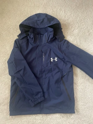 Under Armour mörkblå vind/regn jacka - Stilren svart Under Armour-jacka med riktigt snygg och sportig design. Perfekt för träning, löpning, promenader eller som en clean jacka till vardags. Passas som en stor S och liten M  Jackan har huva, dragkedjefickor och reflexdetaljer som syns tydligt när ljuset träffar dem. Den svarta färgen gör den enkel att matcha med resten av outfiten.  📌 Svart 📌 Under Armour 📌 Sport / träning / löpning 📌 Huva 📌 Reflexdetaljer 📌 Fickor med dragkedja 📌 Snygg och funktionell design  Skickas inom 24 timmar