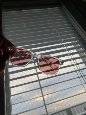 solglasögon med rosa glas - Säljer ett par trendiga solglasögon med transparent båge och rosa tonade glas. Klassisk fyrkantig form som passar till de flesta ansikten. Snygg accessoar för dig som vill sticka ut lite extra i solen.