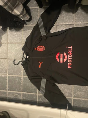Svart AC Milan träningströja  - Snygg svart långärmad träningströja från Puma med AC Milan-logga och röda detaljer. Tröjan har halv dragkedja framtill, gråa ränder på ärmarna och tryck med texten 'FOOTBALL' på bröstet. Perfekt för fotbollsträning eller sport.