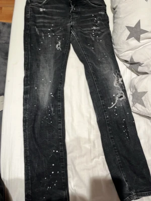 Svarta jeans med slitningar och stänk - Säljer ett par svarta jeans med coola slitningar och vita färgstänk för en edgy look. Jeansen har raka ben och klassisk femficksmodell. Materialet är denim med en något tvättad finish som ger en avslappnad vibe. Dsq2 byxor, där man knäpper byxorna har en knapp gått sönder.