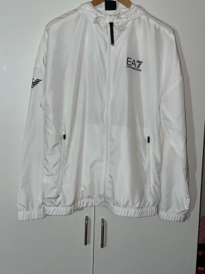 Vit vindjacka från EA7 Emporio Armani - Hej! Säljer en helt ny vit vindjacka från EA7 Emporio Armani med huva. Köpte den ifrån boozt men tyvärr ingenting för mig, qrkod på insidan finns som vissar äktenskapshet🙌