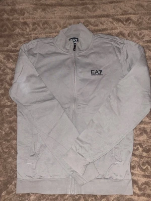 Ljusgrå zip hoodie från Emporio Armani - Snygg ljusgrå zip hoodie från Emporio Armani EA7 med diskret logga på bröstet. Hoodien har dragkedja framtill, ribbade muddar och två fickor. Tillverkad i mjukt bomullsmaterial som känns skönt mot huden. Perfekt för en avslappnad och stilren look.                                                                              Storlek 38 ish?