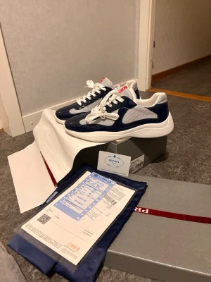 Prada cups  - Snygga sneakers från Prada i marinblått och vitt med meshdetaljer och vita snören. Skorna har en chunky sula och klassisk Prada-logga på plösen. Perfekta för dig som vill ha en stilren och exklusiv look.