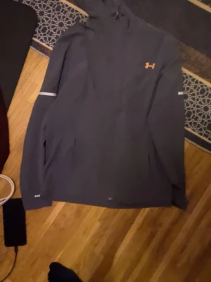 Mörkgrå hoodie från Under Armour - Snygg mörkgrå hoodie från Under Armour med orange logga på bröstet. Hoodien har huva, långa ärmar och reflekterande detaljer på ärmarna. Perfekt för dig som gillar sportig och clean stil.