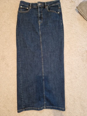 Mörkblå jeanskjol med slits bak - Rak, lång jeanskjol i mörkblå denim med klassiska fem fickor och slits baktill. Kjolen har bälteshällor och kontrastsömmar för en tidlös look. Perfekt för dig som gillar enkel och stilren denim.