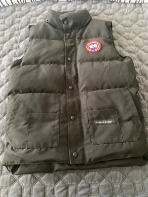 Svart dunväst från Canada Goose - Snygg svart dunväst från Canada Goose med klassisk logga på bröstet och två stora fickor framtill. Västen har hög krage och knäppning med tryckknappar. Perfekt för lager-på-lager och håller dig varm under kyliga dagar.