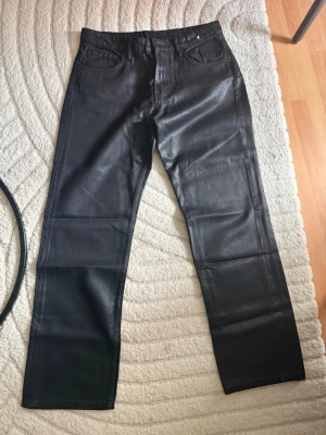 Svarta coated raka jeans - Säljer ett par svarta coated jeans med rak passform och klassisk femficksdesign. Materialet har en glansig finish som ger en cool läderlook. Perfekta för dig som vill ha en edgy stil till din outfit.