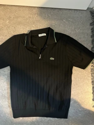 Svart ribbad polotopp från Lacoste - Svart ribbad polotopp från Lacoste med kort ärm och dragkedja framtill. Klassisk krage med diskreta gröna detaljer och den ikoniska broderade krokodilloggan på bröstet. Perfekt för en sportig och stilren look.