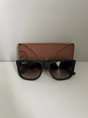 Svarta Ray-Ban solglasögon - Snygga svarta solglasögon från Ray-Ban med fyrkantig form och bruna tonade glas. Kommer med ett klassiskt brunt Ray-Ban-fodral. Perfekta för soliga dagar och ger en cool vibe till din outfit.