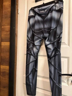 Svarta tights med grafiskt mönster - Säljer ett par svarta tights med grått grafiskt mönster och vita detaljer. Tightsen har hög midja och är tillverkade i ett stretchigt material som sitter tajt mot kroppen. Perfekta för dig som vill ha en unik look under kläderna.