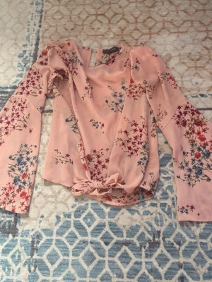 Rosa blommig blus med knytband - Supersöt rosa blus med blommönster. Blusen har långa, vida ärmar och ett knytband nedtill som ger en extra detalj. 