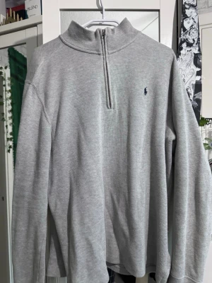 Grå stickad tröja med halvzip Polo - Snygg grå stickad tröja från Polo Ralph Lauren med halv dragkedja och hög krage. Tröjan har lång ärm och klassisk broderad logga på bröstet. Perfekt för lager-på-lager och enkel att matcha med det mesta. Motsvarar M, möjligen L också