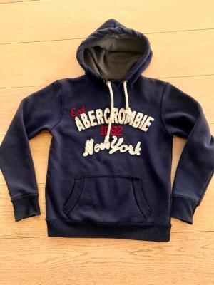 Marinblå hoodie Abercrombie New York - Snygg marinblå hoodie från Abercrombie med broderad text 'Abercrombie 1892 New York' i vitt och rött på bröstet. Hoodien har en stor huva med vita dragsnören och en klassisk magficka. Perfekt för en avslappnad och trendig stil.