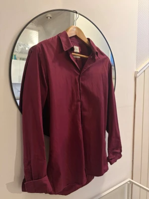 Vinröd bläck skjorta - Snygg vinröd bläck skjorta med klassisk krage och långa ärmar. Skjortan har knappar framtill och är tillverkad i ett mjukt tyg som ger en avslappnad men stilren look. Perfekt att styla till jeans eller kostymbyxor.