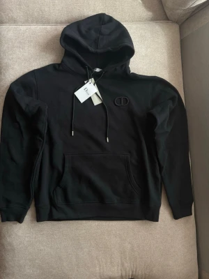 Svart hoodie med CD-logga - Snygg svart hoodie med huva och dragsnören. På bröstet sitter en diskret CD-logga i svart. Hoodien har en stor magficka och ribbade muddar vid ärmar och nederkant. Perfekt för en avslappnad och stilren look. Den är även oversized  så den sitter mer som en L