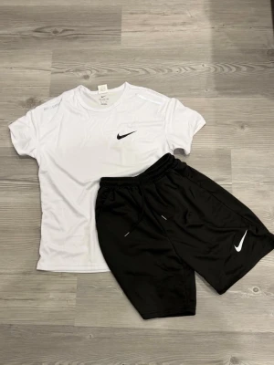 Nike träningsset t-shirt och shorts - Säljer ett sportigt träningsset från Nike med en vit t-shirt och svarta shorts. T-shirten har rund hals och Nike-logga på bröstet, shortsen har resår i midjan och logga på benet. Perfekt för gym eller löpning. Materialet är lätt och andas.
