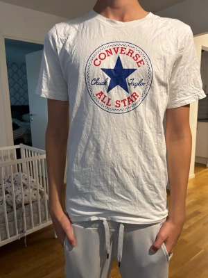 Vit Converse All Star t-shirt - Säljer en vit t-shirt från Converse med det klassiska All Star-trycket i blått och rött framtill. T-shirten har rund hals och korta ärmar. Perfekt för dig som gillar en enkel och sportig stil.