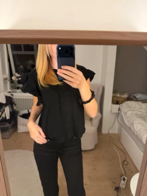 Svart tröja med volangärmar - Svart tröja med korta volangärmar och rund hals. Modellen är något croppad och har en lös passform som ger en avslappnad men trendig look. Perfekt att styla med jeans eller kjol för en cool outfit.