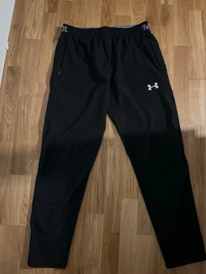 Svarta träningsbyxor från Under Armour - Säljer ett par svarta träningsbyxor från Under Armour med elastisk midja och logga på vänster ben. Byxorna har en smal passform och är perfekta för gymmet eller löprundan. Materialet är lätt och andas bra.