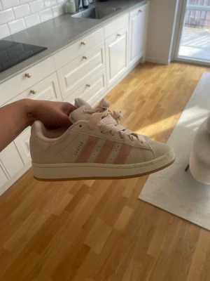 Adidas Campus ljusrosa sneakers - Snygga Adidas Campus sneakers i ljusrosa mocka med klassiska tre ränder i rosa nyans. Skorna har vita snören, låg profil och gummisula. Perfekta för dig som gillar en clean och stilren look med en touch av färg. Finns små fläckar på skorna , se bilder. Använda endast 3 ggr 
