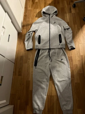 Nike tech tracksuit grå - Snygga gråa mjukisbyxor från Nike med svarta detaljer och dragsko i midjan. Byxorna har muddar vid bensluten och två fickor med dragkedja. Perfekta för chill eller träning, riktigt sköna och stilrena.