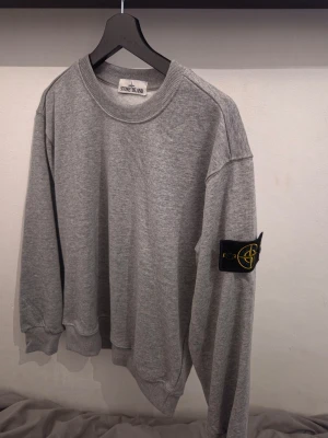 Stone island grå - Klassisk grå sweatshirt från Stone Island med rund halsringning och ribbade muddar. Tröjan har den ikoniska Stone Island-loggan på vänster ärm och är tillverkad i mjukt bomullsmaterial. Perfekt för en avslappnad och stilren look.