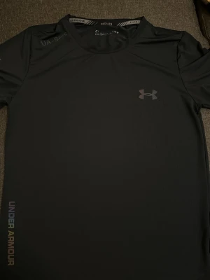 Svart träningströja Under Armour - Snygg svart kortärmad träningströja från Under Armour med diskret logga på bröstet och tryck på sidan. Tillverkad i ett lätt och snabbtorkande funktionsmaterial som passar perfekt till gymmet eller löpning.