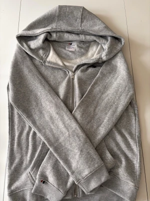 Grå hoodie från U.S. Polo Assn. - Snygg grå hoodie från U.S. Polo Assn. med dragkedja framtill och två fickor. Tillverkad i mjukt sweatshirtmaterial och har en klassisk huva. Perfekt för en avslappnad och sportig stil.