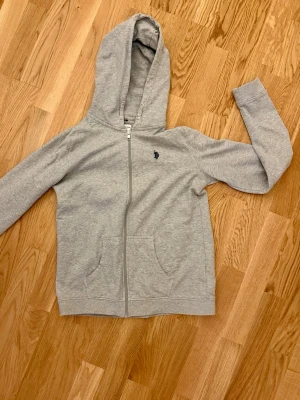 Grå hoodie från U.S. Polo Assn. Knappt använd!  - Snygg grå hoodie från U.S. Polo Assn. med dragkedja framtill och två fickor. Klassisk logga broderad på bröstet och stor huva. Tillverkad i mjukt sweatshirtmaterial som är skönt att ha på sig. Perfekt för chill dagar eller när du vill ha en enkel och clean look. Storleken är 15-16 och motsvarar 164 i barn. 